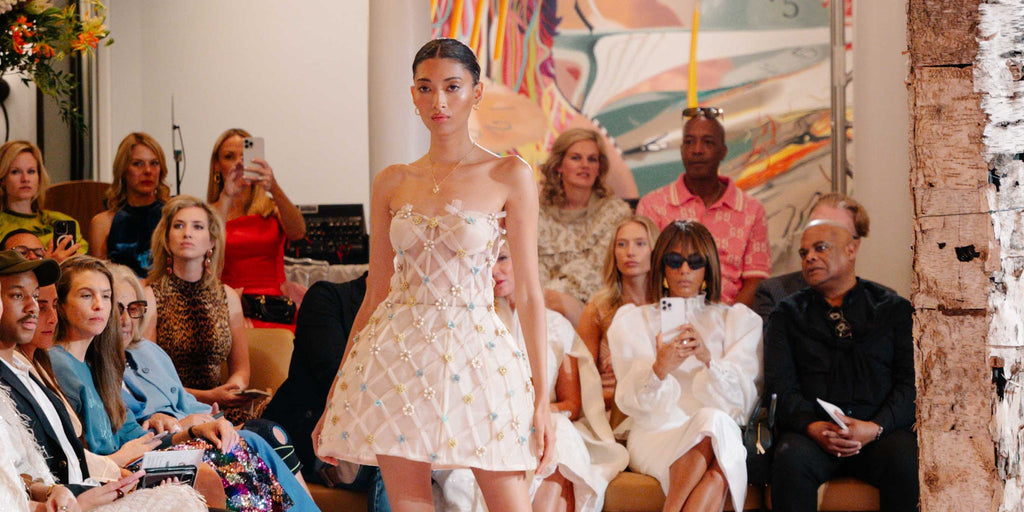 Model in a strapless embellished mini dress walks the runway at an indoor fashion show, with a stylish audience watching and capturing the moment.